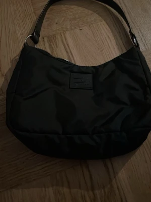 Svart axelväska i syntetmaterial - Snygg svart axelväska från NA-KD i mjukt syntetmaterial. Väskan har en enkel, stilren design med en bred axelrem och dragkedja upptill. Perfekt storlek för det viktigaste och passar till många olika outfits.