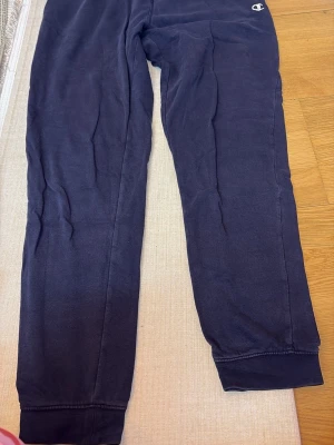 Mörkblå mjukisbyxor från Champion - Säljer ett par mörkblå mjukisbyxor från Champion med resår i bensluten och en liten logga vid höften. Byxorna är tillverkade i mjukt bomullsmaterial och har en avslappnad passform som är perfekt för chill dagar.
