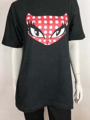 Svart t-shirt med kattmotiv och prickar storlek L. - Cool svart t-shirt med ett stort tryck av en katt med röda och vita prickar och intensiva ögon på bröstet. T-shirten har korta ärmar och en relaxed passform. Perfekt för dig som gillar statement-plagg och grafiska prints. Storlek L. Helt ny.