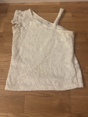 Vit spets topp oneshoulder - Säljer en vit topp i spets med blommigt mönster. Toppen har oneshoulder-design med en axelbandslös sida och en axel med spetsdetalj. Perfekt för dig som gillar romantisk och unik stil.