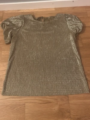 Glittrig guld blus med puffärm - Säljer en glittrig guldblus med korta puffärmar och rund hals. Tyget har en ribbad struktur som ger extra effekt och blusen stängs med en knapp i nacken. Perfekt för dig som vill sticka ut lite extra.
