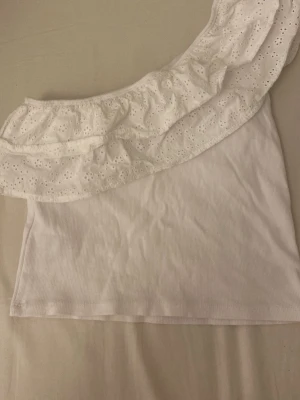 Vit oneshoulder topp från Gina Tricot - Säljer en vit oneshoulder topp från Gina Tricot i ribbad bomull. Toppen har en bred volang med broderade hål och är axelbandslös på ena sidan.  Använd 1 gång bara🩷