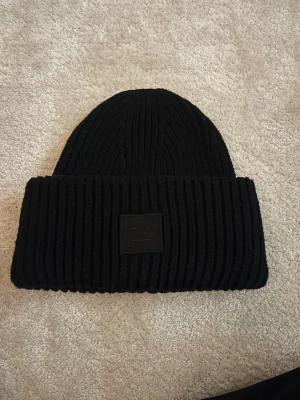 Mössa Acne Studios - Säljer en svart ribbstickad mössa från Acne Studios med den klassiska ansiktsloggan framtill. Mössan är i tjockt, mjukt material och har en bred uppvikt kant. Perfekt för kalla dagar och ger en clean, stilren look.