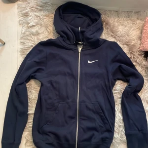 Mörkblå Nike hoodie med dragkedja XS - Snygg mörkblå hoodie från Nike med vit logga på bröstet. Tröjan har huva med snörning, dragkedja framtill och två fickor. Perfekt för chill dagar eller när du vill ha en sportig look.