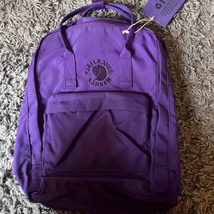 Lila Fjällräven Kånken ryggsäck - Säljer en lila Fjällräven Kånken ryggsäck med klassisk fyrkantig form och broderad logga framtill. Väskan har justerbara axelremmar, topphandtag och en praktisk framficka med dragkedja. Materialet är slitstark polyester som tål vardagens äventyr. Nyskick med prislapp 