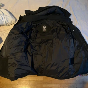 Svart dunjacka från Canada Goose - Säljer en svart dunjacka från Canada Goose med klassisk logga på ärmen. Jackan är tjock och varm, perfekt för kalla dagar. Medföljer originalpåse och avtagbar pälskant till huvan. Jackan har dragkedja och hög krage för extra skydd mot kyla.