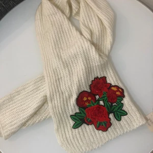Vit stickad halsduk med ros - FRI FRAKT - Snygg vit stickad halsduk från Viritosi med broderade röda blommor och gröna blad som detalj. Halsduken är mjuk och har en grovstickad struktur som ger en cool och mysig vibe. Perfekt för att lyfta din vinterstil med lite färg.