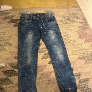 Blå jeansbyxor med snörning från Sarol - Säljer ett par blå jeansbyxor från Sarol med resår och snörning i midjan. Byxorna har en avslappnad passform, elastiska benslut och klassiska fickor fram och bak. Perfekta för en chill och bekväm stil.