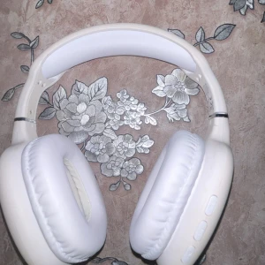 Trådlösa over-ear hörlurar, vit - Stilrena vita trådlösa over-ear hörlurar med mjuka öronkuddar och justerbar huvudbygel. Flera knappar för volym och uppspelning på sidan. Hörlurarna ser ut att vara i gott skick utan synligt slitage eller defekter. Perfekt för musik, film och samtal.