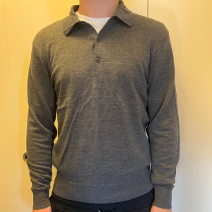 Grå pikétröja från Uniqlo - Snygg grå långärmad pikétröja från Uniqlo med klassisk krage och två knappar framtill. Tröjan är 100% ull och har en normal passform som sitter skönt. Perfekt för dig som gillar stilrena och enkla plagg. Köparen betalar frakt!