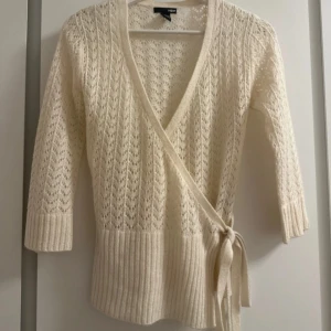 Beige omlottkofta från H&M - Superfin beige stickad omlottkofta från H&M med knyt i sidan och trekvartsärm. Koftan har ett snyggt hålmönster och bred ribbad kant nedtill och vid ärmslut. Perfekt för lager-på-lager och ger en chill men ändå stilren vibe. Passar på xs-s