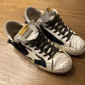 Golden Goose sneakers med stjärndetalj - Golden Goose sneakers i vit och svart med ikonisk stjärna på sidan. Ovandel i skinn och textil med slitna detaljer och svarta snören. Gula och vita detaljer på hälen och tungan. Snygg vintage-look och coola kontraster. Storlek - 42 