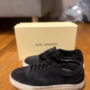 Svarta sneakers från Axel Arigato - Snygga svarta sneakers från Axel Arigato med mockaöverdel och vit platt sula. Skorna har rund tå och klassisk snörning. Perfekt för dig som gillar stilrena och minimalistiska sneakers med exklusiv känsla.