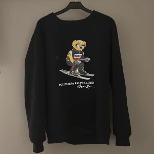 Svart sweatshirt Polo Bear Ralph Lauren - Svart sweatshirt från Polo Ralph Lauren med ikoniskt Polo Bear-tryck där björnen åker skidor.Storlek L men passar M med. Tröjan har rund halsringning och långa ärmar. Perfekt statement-piece för dig som gillar streetwear och sportiga vibes.