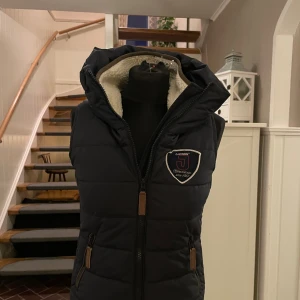 Svart dunväst från Jacson med krage - Säljer en svart dunväst från Jacson med mysigt teddyfoder i kragen och två dragkedjefickor. Västen har en logga på bröstet och hög krage som skyddar mot vind. Perfekt för dig som gillar ridning eller vill ha en varm väst till höst och vinter. Säljer den pågrund av att den blivit för liten. Använt den väldigt få gånger.🤗