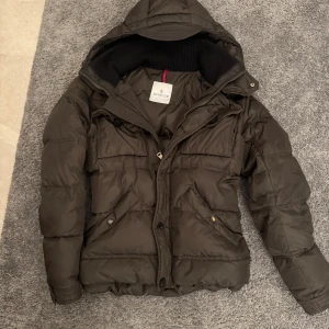 Moncler pufferjacka - Säljer en svart pufferjacka från Moncler med huva och dragkedja. Jackan har en quiltad design, två fickor framtill med knappar och ribbade muddar vid ärmslut. Perfekt för kalla dagar, en knapp på jackan är lite sönder (se sista bild)