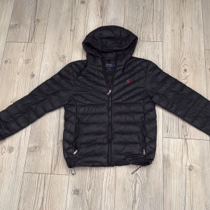 Svart dunjacka från Polo Ralph Lauren - En riktigt fet svart dunjacka från Polo Ralph Lauren med huva och dragkedja framtill. Jackan har två fickor med dragkedja och ikonisk röd broderad logga på bröstet. Lätt quiltad design och justerbar dragsko nedtill. Perfekt för kyliga dagar. Nypris 2500.
