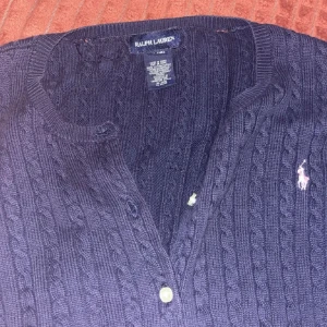 Mörkblå kabelstickad kofta Ralph Lauren - Kabelstickad mörkblå kofta från Ralph Lauren med klassisk passform. Koftan har långa ärmar, rund halsringning och knäppning framtill med vita knappar. Ikoniska broderade loggan i vitt på bröstet. Perfekt för lager-på-lager-stil.