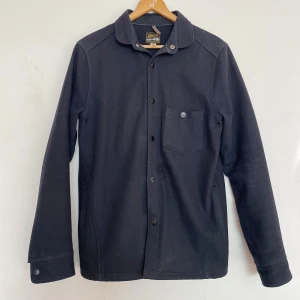 Mörkblå overshirt från Sarva - Stilren mörkblå overshirt från Sarva med klassisk krage, knappstängning och en bröstficka med knapp. Jackan är tillverkad i ullblandning och har långa ärmar samt diskreta sömdetaljer. Perfekt lager-på-lager-plagg för kyligare dagar.
