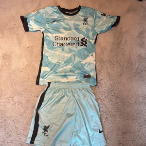 Liverpool M. Salah fotbollströja blå - Säljer en Liverpool fotbollströja med M. Salah och nummer 11 på ryggen. Tröjan är ljusblå med ett unikt camouflage-mönster och svarta detaljer. Märkt med Nike-logga och LFC-emblem. Materialet är lätt och andas, perfekt för match eller träning.