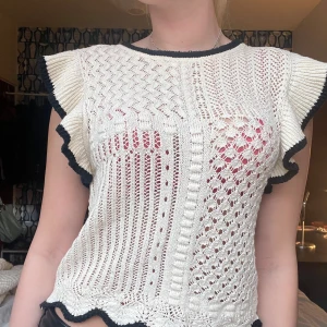 Vit stickad blus från Zara med volang - Superfin vit stickad blus från Zara med svarta kanter och volangärmar. Blusen har ett genombrutet mönster i olika stickade strukturer och vågig nederkant. Perfekt för dig som gillar unika detaljer och en söt, trendig look.