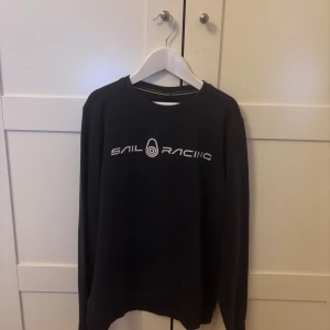 Svart Sail Racing sweatshirt - Svart sweatshirt från Sail Racing med vit logga tryckt på bröstet och en mindre logga i nacken. Klassisk rund halsringning. Märkena har slitits lite av tvättmaskinen. Den har dock används mycket väl. Kan sänka priset.