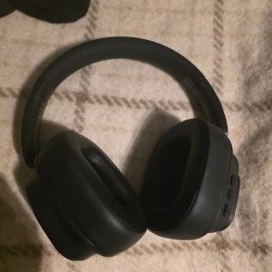 Trådlösa over-ear hörlurar med inbyggd display - Stilrena svarta trådlösa over-ear hörlurar med mjuka kuddar och inbyggd färgdisplay som visar tid och batterinivå. Flera knappar för enkel styrning. Hörlurarna ser ut att vara i mycket gott skick utan synligt slitage. Perfekt för musik, samtal och vardagsbruk.