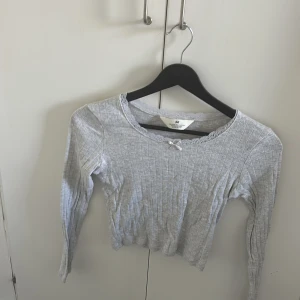 Ljusgrå ribbad topp med rosett H&M - Supersöt ljusgrå ribbad topp från H&M med lång ärm och liten rosett vid halsringningen. Croppad modell med diskret spetskant runt halsen. Perfekt för dig som gillar en enkel men gullig stil.