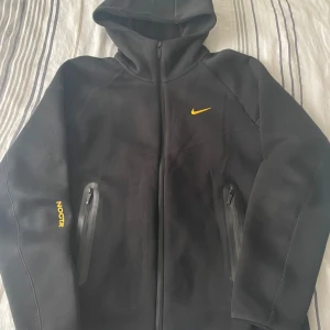 Nike NOCTA svart tracksuit med huva - Svart tracksuit från Nike NOCTA med gul logga på både jacka och byxa. Jackan har huva, hel dragkedja och två fickor med dragkedja. Byxorna har snörning i midjan och dragkedjeficka. Snygg och sportig look med NOCTA-tryck i gult på ärm och ben.
