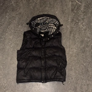 Svart dunväst från Burberry - Svart dunväst från Burberry med avtagbar huva och klassiskt rutigt foder i grått, vitt och svart. Västen har dragkedja, knappar och två fickor framtill. Perfekt för lager-på-lager och snygg streetstil. Stolek L. 