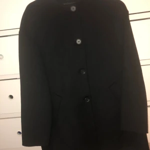 Svart klassisk ullkappa - Stilren svart kappa i ull med rak passform och knäppning framtill med fyra knappar. Jackan har diskreta fickor på sidorna och en minimalistisk design som passar perfekt till höst och vinter. Enkel och tidlös modell som är lätt att matcha.