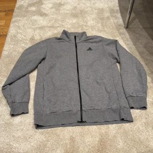 Grå ziptröja från Adidas - Snygg grå ziptröja från Adidas med svart logga på bröstet och stor adidas-tryck på ryggen. Tröjan har hel dragkedja, ståkrage och ribbade muddar. Perfekt för träning eller chill. Materialet är mjukt och skönt, troligtvis bomullsmix.