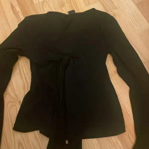 Svart långärmad topp med knyt - Snygg svart långärmad topp med knyt framtill som ger en cool och trendig look. Toppen har en figurnära passform och är perfekt att styla med jeans eller kjol. Enkel men ändå med en twist tack vare knytet.