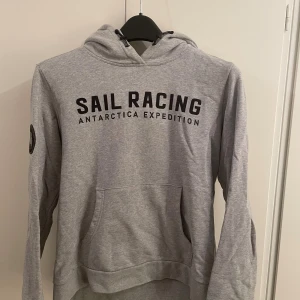 Grå hoodie från Sail Racing - Snygg grå hoodie från Sail Racing med svart tryck på bröstet och coolt expeditionsmärke på ärmen. Tröjan har huva med snörning och en stor magficka. Perfekt för dig som gillar sportig och avslappnad stil.