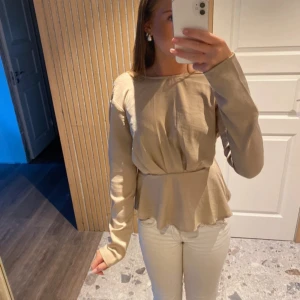 Beige blus med öppen rygg och volang - Säljer en beige blus med lång ärm och snygg volang nertill. Toppen har en markerad midja och en djup, öppen rygg som ger en cool touch. Perfekt för dig som gillar stilrena och trendiga plagg med det lilla extra.