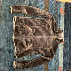  fleece från Jacson - Mysig fleecejacka från Jacson med dragkedja framtill och två fickor med snygga läderdetaljer. Jackan har hög krage och är helfodrad i mjuk fleece. Perfekt för kyliga dagar när du vill vara både varm och bekväm. Storlek 158/164. Liten defekt på en av läderdetaljerna.