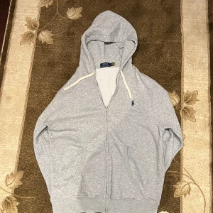 Grå hoodie från Polo Ralph Lauren - Klassisk ljusgrå hoodie från Polo Ralph Lauren med dragkedja, huva med vita snören och broderad blå logga på bröstet. Mjuk bomullskänsla och två fickor framtill. Perfekt för chill dagar eller när du vill ha en enkel och stilren look. Helt oanvänd!
