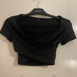 Svart omlott croppad topp från SHEIN - Säljer en svart croppad topp från SHEIN med omlott-design och korta ärmar. Toppen har en snygg v-ringning och sitter tight mot kroppen. Perfekt för dig som gillar stilrena och trendiga plagg med enkel look.