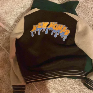 Grön och brun bomberjacka från Woodbird - Säljer en fet bomberjacka från Woodbird med grön och brun kropp, beige ärmar och coola broderade detaljer. Jackan har tryckknappar, randiga muddar och tryck på både fram- och baksida med texten 'New Nomad'. Perfekt statement-plagg för streetwear. 1200kr kosta den ny knappt använd priset kan sänkas 