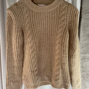 Beige stickad tröja från KIDS ONLY - Mysig beige stickad tröja från KIDS ONLY med grovstickat mönster och flätade detaljer framtill. Tröjan har rund halsringning och långa ärmar, perfekt för kyliga dagar. Enkel att matcha med jeans eller kjol.