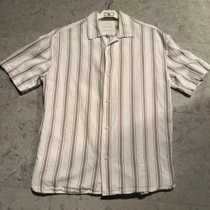 Skjorta - Snygg kortärmad skjorta från Jack & Jones i vitt med beige och bruna ränder. Skjortan har klassisk krage och knappar framtill. Perfekt för dig som gillar en chill och stilren look. Materialet är mjuk bomull som känns skönt mot huden.