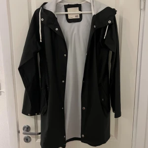 Svart regnjacka med huva från 157 - Svart regnjacka från 157 med vit insida och huva med dragsko. Jackan har tryckknappar framtill, fickor med lock och en stilren, minimalistisk look. Perfekt för regniga dagar och enkel att matcha med allt.