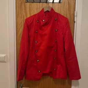 Röd skepparkavaj med dekorativa knappar - En snygg röd skepparkavaj dekorativa svarta knappar med silverfärgade emblem. Jackan har hög krage och raka fickor framtill. Passformen är normal och materialet känns som polyester. Perfekt för maskerader av olika slag och dig som söker en klassisk uniform. Skickas ca 1-3 dagar efter köp. 