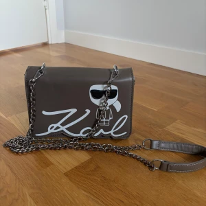 Brun axelväska med kedja - Cool brun axelväska med silverfärgad kedjerem och ikoniskt Karl-tryck framtill. Inte äkta, fick den. Kedjan har tappat färg på några ställen.