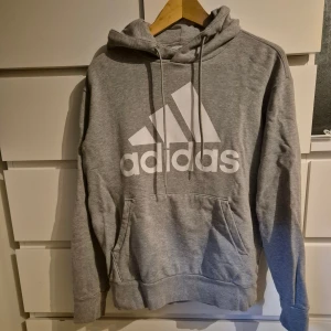 Grå Adidas hoodie med logga - Snygg grå hoodie från Adidas med stor vit logga framtill. Klassisk känguruficka, dragsko i huvan och mjukt material. Perfekt för chill eller träning. Långärmad och normal passform.