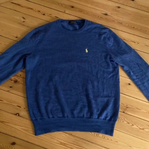 Blå stickad tröja från Polo Ralph Lauren - Klassisk blå stickad tröja från Polo Ralph Lauren med rund halsringning och gul broderad logga på bröstet. Tröjan har ribbade muddar vid ärmslut och nederkant samt lång ärm. Perfekt för dig som gillar stilren och tidlös design.