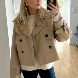 Beige kort trenchcoat - Snygg beige kort trenchcoat med dubbelknäppning och bältdetaljer vid ärmsluten. Perfekt för en stilren look. Jackan har en klassisk krage och är i en tidlös design.