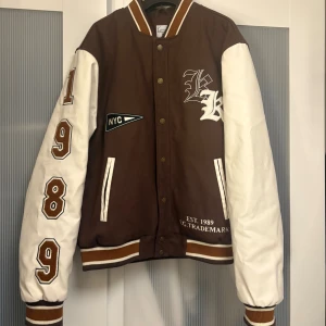 Brun och vit bomberjacka Karl Kani S - Snygg bomberjacka från Karl Kani i brunt och vitt med broderade detaljer, stora siffror på ärmen och coola NYC-patchar. Jackan har ribbade muddar, tryckknappar och kontrastfärgade ärmar. Perfekt för dig som vill sticka ut med streetstyle. Nypris 1500kr