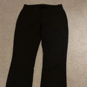 Svarta kostymbyxor från BikBok, stl 36 - Svarta kostymbyxor från BikBok i klassisk modell med raka ben och diskret slits nertill. Snygg passform och stilren look, perfekta till en trendig outfit. Byxorna är i ett mjukt och följsamt material som känns bekvämt hela dagen. Inga noppror eller liknande