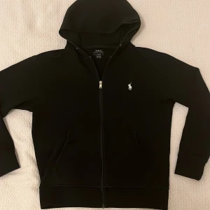 Svart hoodie från Polo Ralph Lauren - Svart hoodie från Polo Ralph Lauren med dragkedja och klassisk vit logga på bröstet. Tröjan har huva, fickor framtill och är tillverkad i mjukt material. Perfekt för en chill och stilren look.
